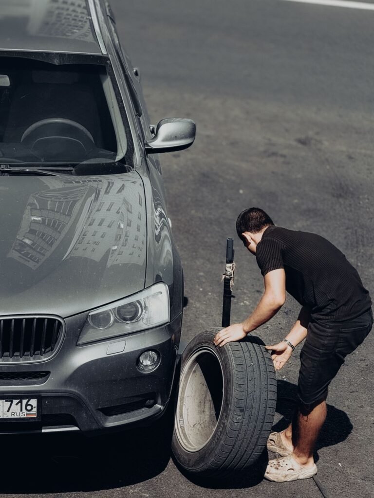 A young man changes a car tire on an outdoor road under bright sunlight